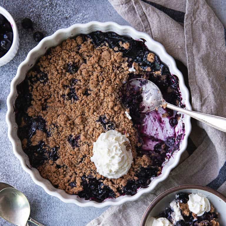 Overhead view of blueberry crumble with a portion scooped out to show the jammy berry filling, topped with whipped cream.